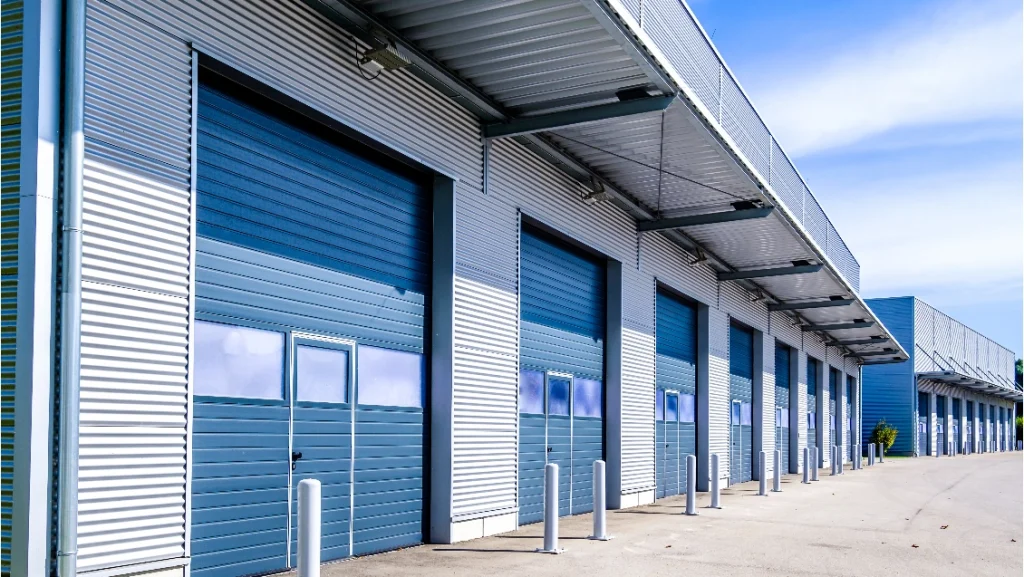 A long, grey steel storage building with a series of blue shutter doors on the front, pictured in front of a blue sky.