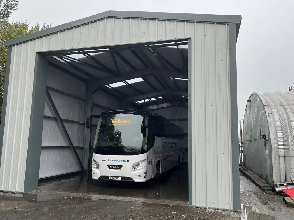 Grey industrial steel building with doors open and large white coach parked inside.