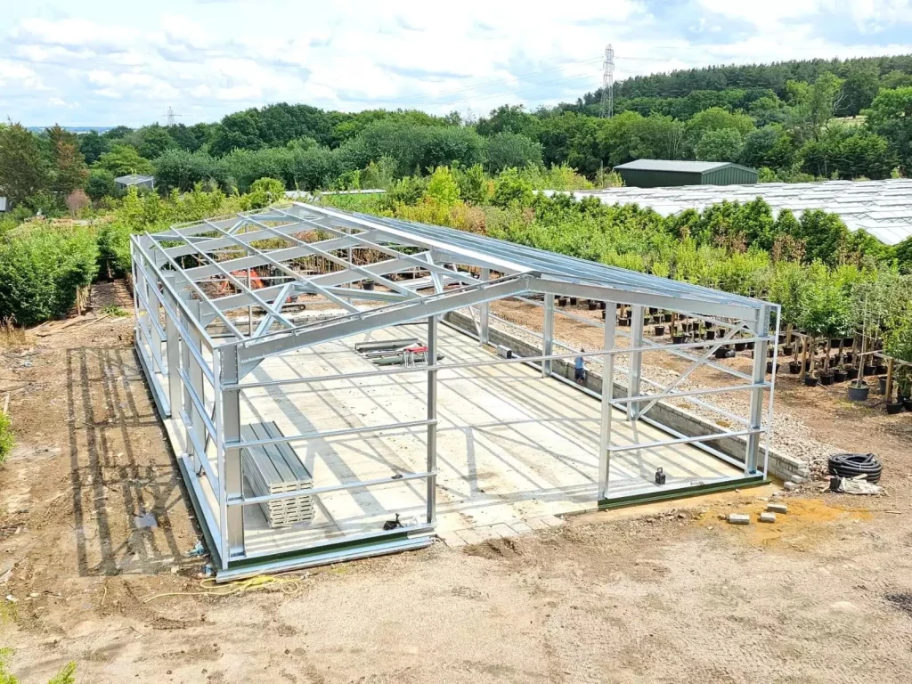 Steel frame building under construction, showing completed grey cladding on one side, exposed steel frame on the other, and brick pillars at the front.