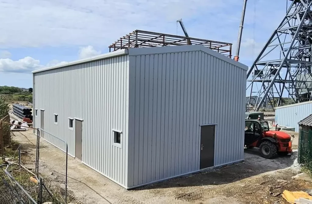 Grey clad steel building at South Crofty Mine with the mine headframe and further steelwork behind