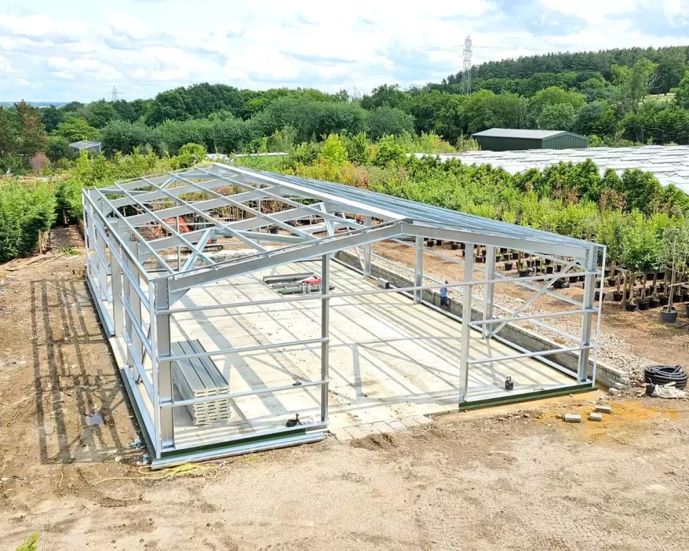 Steel frame building under construction, showing completed grey cladding on one side, exposed steel frame on the other, and brick pillars at the front.