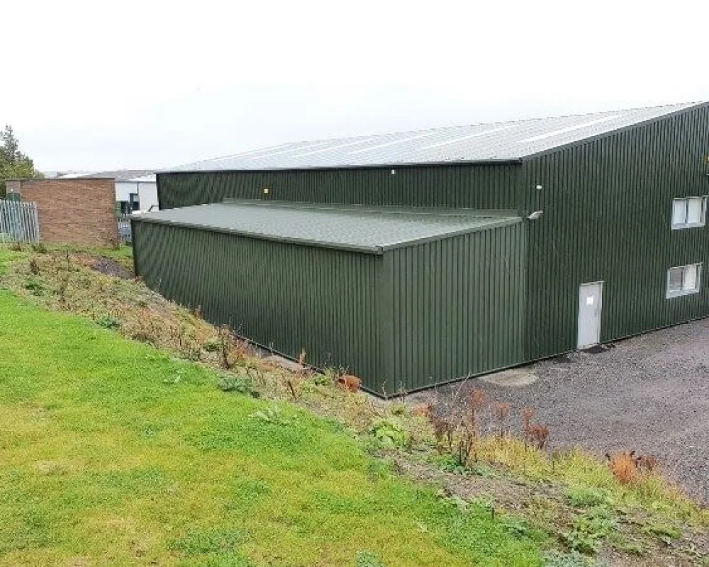 A large green steel building with a steel frame extension attached, viewed from a slightly elevated angle.