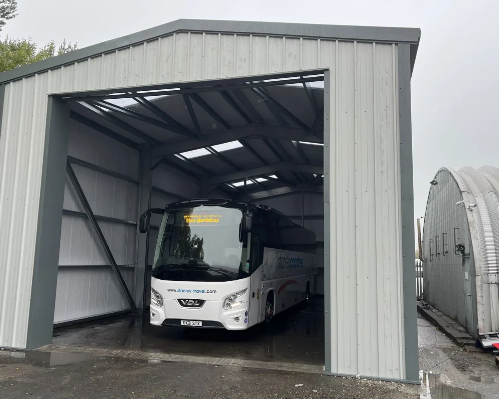Grey industrial steel building with doors open and large white coach parked inside.