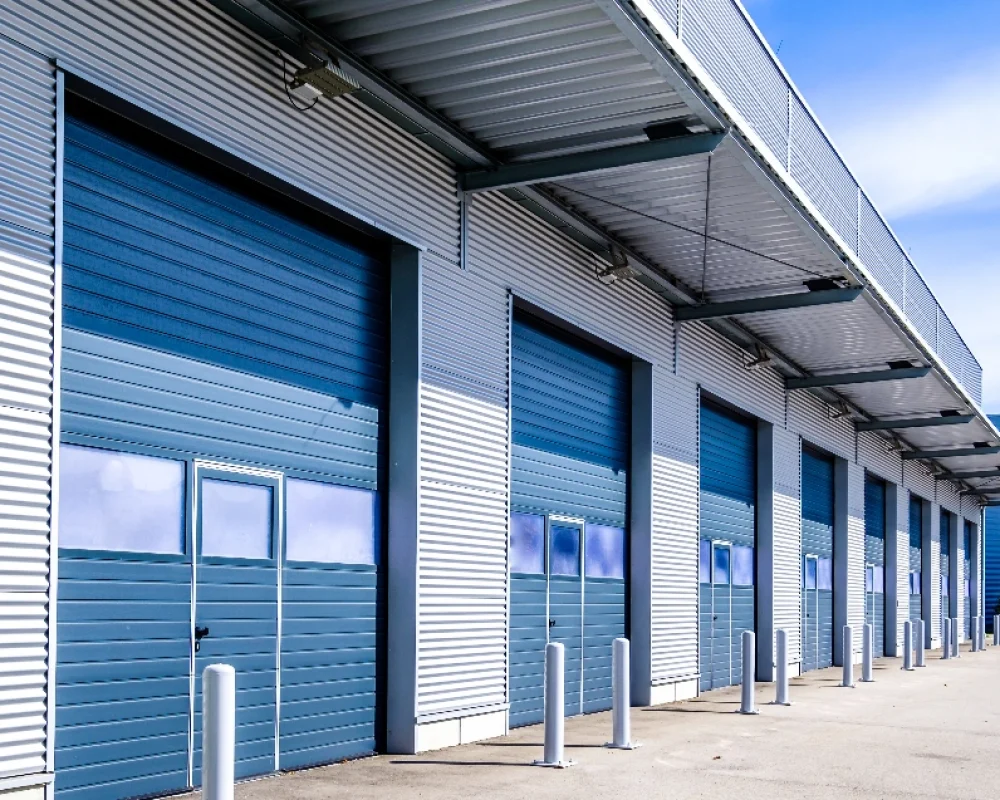 A long, grey steel storage building with a series of blue shutter doors on the front, pictured in front of a blue sky.